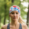 A woman jogging outdoors on a tree-lined path, wearing a white athletic headband with a colorful red, blue, and green floral pattern and a gray tank top.