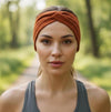 A woman wearing a rust-orange twisted headband and a dark gray athletic tank top, standing on a blurred forest path.