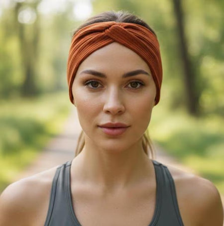 A woman wearing a rust-orange twisted headband and a dark gray athletic tank top, standing on a blurred forest path.