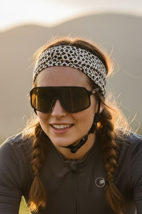 Woman riding a bicycle on a scenic road with mountains in the background with a cheetah headband on