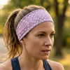 A woman wearing a dusty purple athletic headband with a white leaf pattern, shown in a close-up outdoor shot with a blurred forest background.