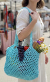 A woman in a white dress carries a large turquoise crochet mesh shoulder bag filled with grapes, a tomato, bread, and flowers in an outdoor market setting.