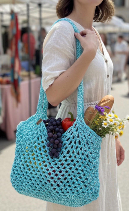 A woman in a white dress carries a large turquoise crochet mesh shoulder bag filled with grapes, a tomato, bread, and flowers in an outdoor market setting.