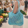 A woman at an outdoor farmers market carries a large, light blue crocheted mesh tote bag over her shoulder while holding a green vegetable.