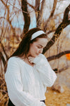 Woman wearing a white cable knit cardigan and white workout headband standing outdoors by tree branches