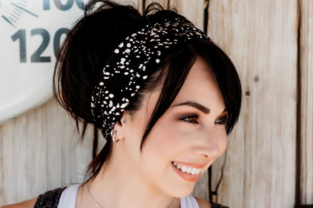 Smiling woman wearing a black and white speckled workout headband with a white tank top and pendant necklace against rustic wood background