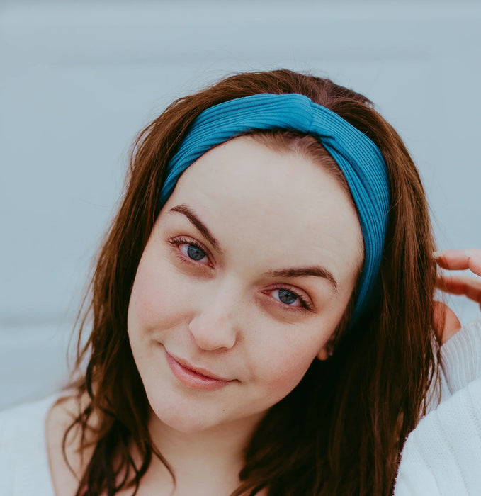Young woman wearing a handmade blue workout headband and white sweater standing against a light gray background