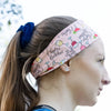 Side profile of woman wearing pink workout headband with colorful drinks pattern and text, blue tank top outdoors
