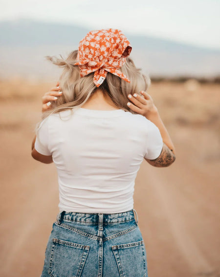 Woman in white t-shirt and high-waisted jeans wearing an orange floral workout hair scarf outdoors on a dirt path