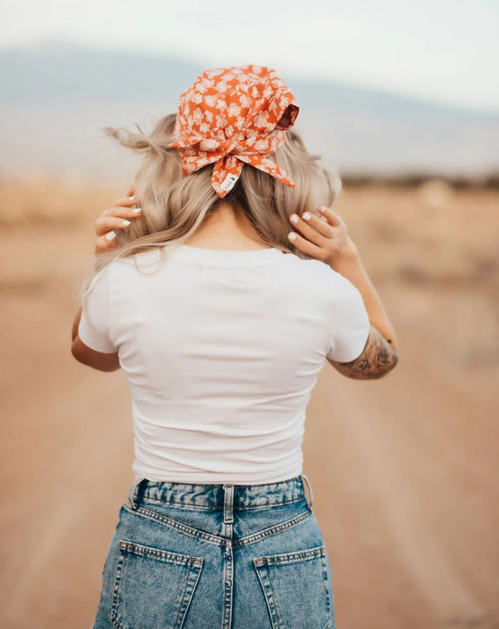Woman in white t-shirt and high-waisted jeans wearing an orange floral workout hair scarf outdoors on a dirt path