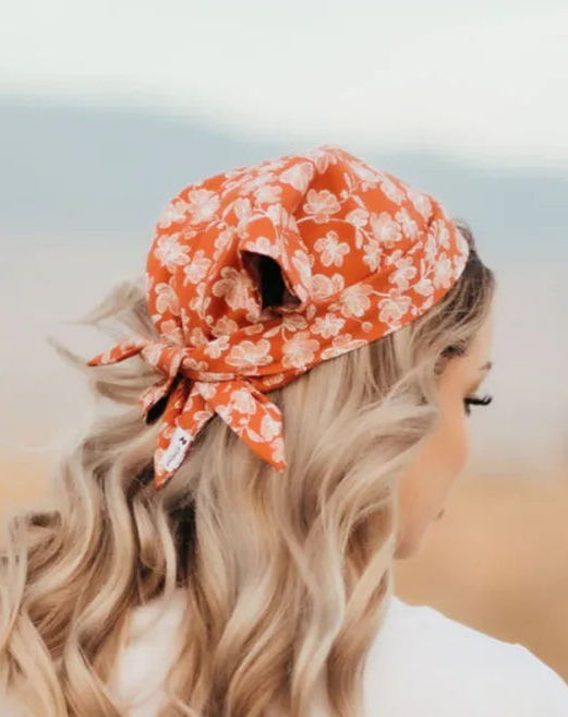 Woman with long blonde hair wearing an orange floral workout hair scarf, white t-shirt, and denim shorts outdoors