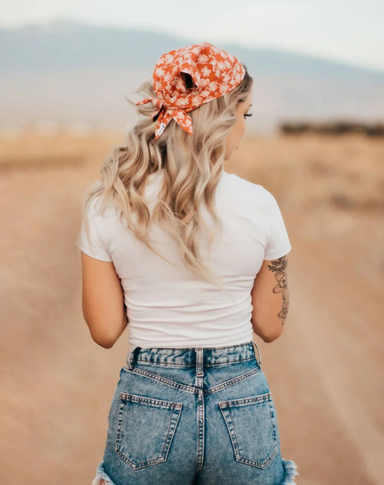 Woman with long blonde hair wearing an orange floral workout hair scarf, white t-shirt, and denim shorts outdoors