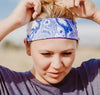 Woman outdoors adjusting blue and white handmade workout headband with Greek Isles pattern, wearing purple t-shirt