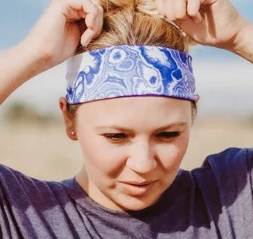Woman outdoors adjusting blue and white handmade workout headband with Greek Isles pattern, wearing purple t-shirt