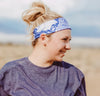 Woman wearing a blue and white patterned workout headband and purple shirt smiling outdoors at a beach setting