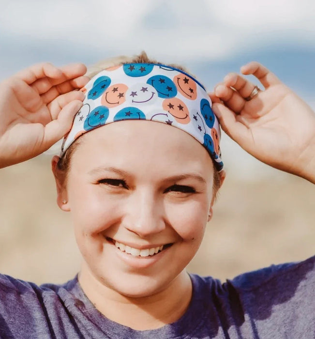 Smiling woman adjusting colorful smiley face workout headband outdoors, wearing a purple athletic shirt
